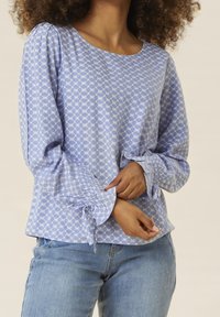 Blusa blu chiaro con stampa geometrica bianca, maniche lunghe con polsini arricciati, scollatura rotonda e tessuto morbido. Abbinata a jeans.