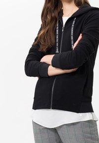 Black zip-up hoodie with a textured design and logo accents on drawstrings, featuring ribbed cuffs, worn over a white shirt and plaid trousers.