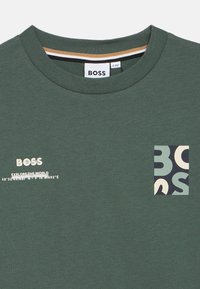 Πράσινη βαμβακερή μπλούζα με στρογγυλή λαιμόκοψη, που φέρει ένα μικρό λογότυπο στο μπροστινό μέρος, το κείμενο "ΕΞΕΡΕΥΝΗΣΤΕ ΤΟΝ ΚΟΣΜΟ" και ένα τσέπη με σχέδιο σε αντίθετα χρώματα και σχήματα.