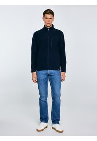 Mężczyzna stojący na jednolitym tle, ubrany w granatową bluzę polarową na zamek, niebieskie dżinsy i białe trampki.