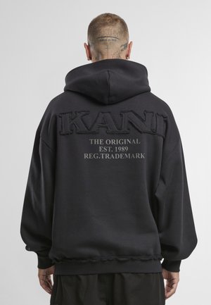 Sort hættetrøje med broderet "KANI"-logo og tekst på ryggen. Materialet virker blødt; designet har en stor hætte og ribbede manchetter.