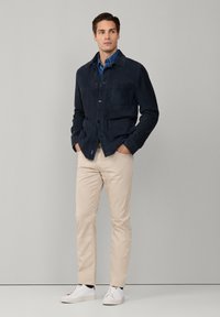 Marineblauwe suède jas met knoopsluiting en twee voorzakken, gecombineerd met beige slim-fit broek en witte sneakers op een neutrale achtergrond.