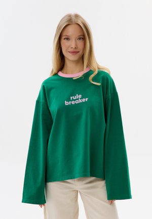 Jeune femme aux longs cheveux blonds portant un sweat-shirt vert oversize avec une bordure rose et le texte « rule breaker », debout devant un fond uni.