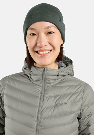 Smiling person wearing a grey beanie, grey padded jacket with hood, and small gold hoop earrings against a plain white background.