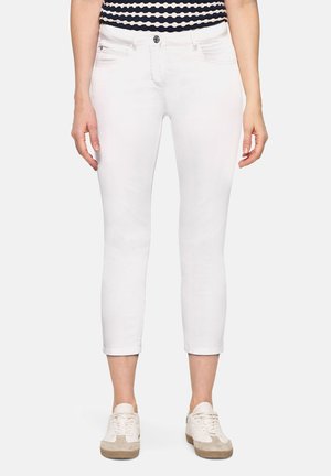 Kvinde iført hvide korte slim-fit bukser, navy blå og hvid mønstret top og hvide sneakers, stående mod en ensfarvet baggrund.