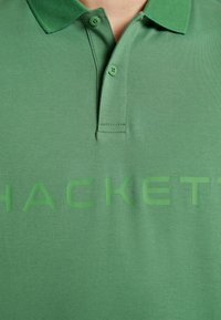 Polo en coton vert avec un col, deux boutons et un grand logo en relief affichant « HACKETT » dans un vert assorti.