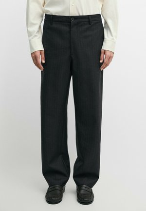 Man wearing black pinstripe trousers, black loafers, and a cream long-sleeve buttoned shirt, standing against a plain background.