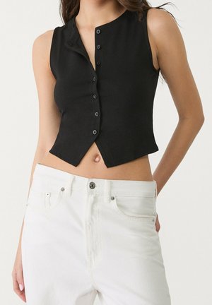 Vrouw die een mouwloze zwarte crop top met knopen en een witte jeans met hoge taille draagt, staand tegen een effen lichte achtergrond.