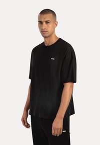 T-shirt nera oversize in cotone con maniche corte, caratterizzata da un piccolo logo sul petto. Il tessuto ha una consistenza liscia e il design è minimalista.