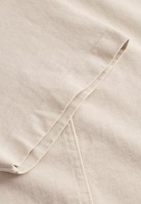 Beiger Baumwollstoff mit glatter Textur, der einen gefalteten Saum und feine Stickdetails entlang der Kante aufweist. Der Stoff ist leicht zerknittert.