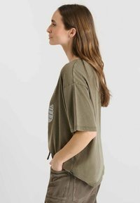 Olijfgroene oversized T-shirt met een ronde hals, korte mouwen en een bedrukking aan de voorkant, gecombineerd met gestructureerde bruine broek.