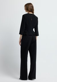 Černý šity peplum top s tříčtvrtečními rukávy a klíčovou dírkou vzadu, v kombinaci s černými kalhotami s vysokým pasem, vytvářející elegantní siluetu.