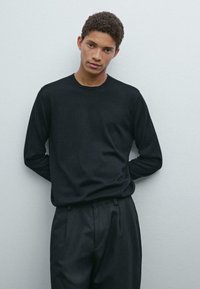 Black long-sleeve crew neck sweater made of smooth fabric, paired with dark tailored trousers. Model stands with hands behind back.