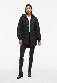 Chaqueta negra acolchada con capucha, que presenta cierres a presión, puños acanalados y un corte relajado. Llevada sobre una camiseta blanca y pantalones vaqueros negros.
