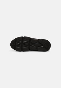 Semelle de chaussure en caoutchouc noir avec motif de bande de roulement géométrique, design segmenté et zones d'adhérence texturées sur un fond clair uni.