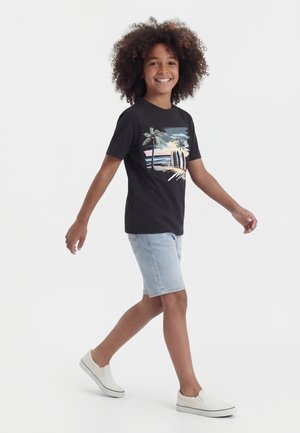Χαμογελαστό παιδί με σγουρά μαλλιά που περπατά, φοράει μαύρο μπλουζάκι με στάμπα φοίνικες και ωκεανό, ανοιχτόχρωμα τζιν σορτς και λευκά slip-on παπούτσια.