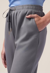 Graue Jogginghosen mit elastischem Bund, Kordelzug mit goldenen Akzenten, seitlicher Tasche und "SOFT LIFE" Textdetail am Bein. Glatte Textur.