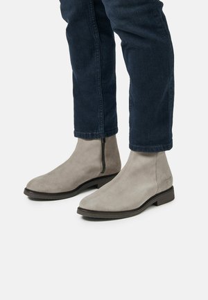 Botas chelsea de ante gris con un diseño elegante, que presentan un cierre lateral y una punta redondeada, combinadas con vaqueros azul oscuro.