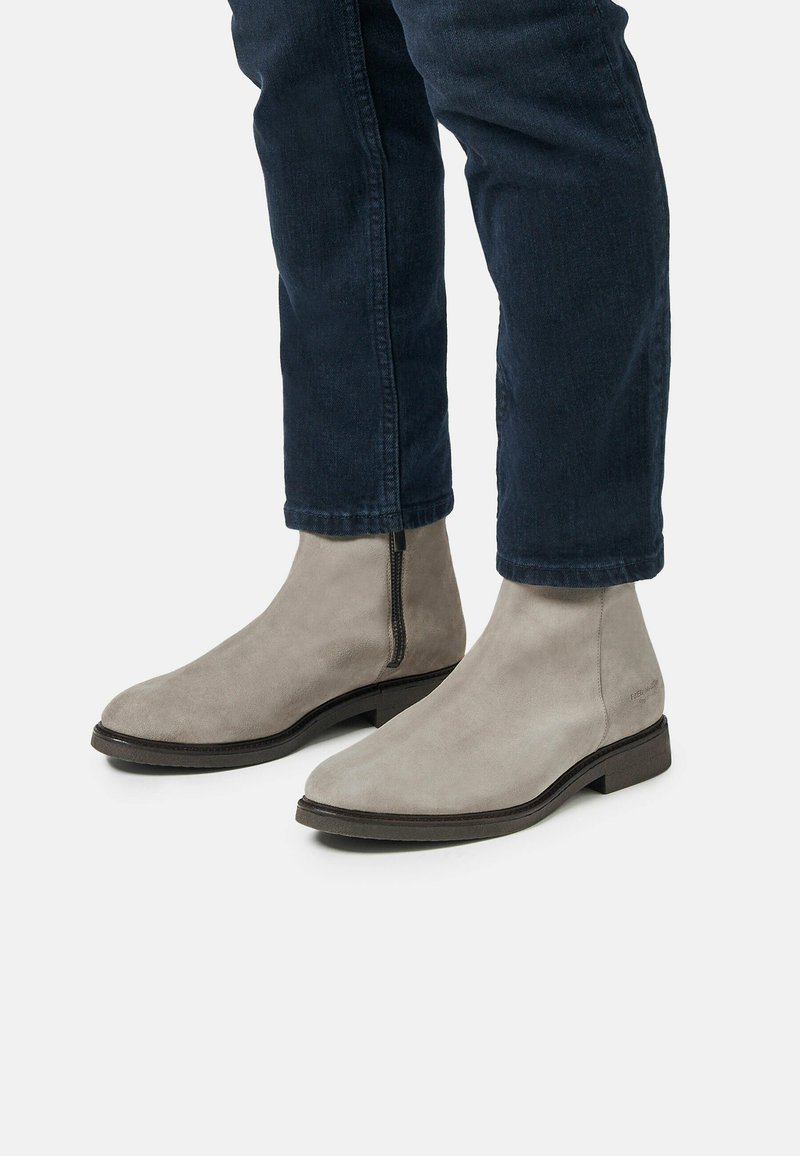 Botas chelsea de ante gris con un diseño elegante, que presentan un cierre lateral y una punta redondeada, combinadas con vaqueros azul oscuro.