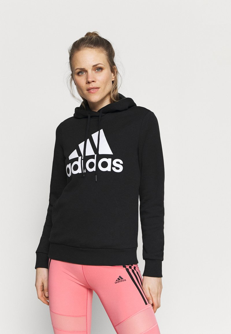 Felpa nera con un grande logo bianco Adidas, dotata di cappuccio con cordino e polsini a costine, abbinata a leggings rosa con strisce nere.