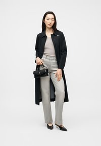 Γυναίκα που φοράει μαύρο μακρύ παλτό, γκρι ριγωτό τοπ, ανοιχτό γκρι τζιν, μαύρες γόβες και κρατάει μια μικρή μαύρη τσάντα.