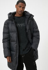 Sort pufferjakke med hætte, der har quiltede sektioner og en blank overflade. Båret over en mørk, struktureret sweatshirt.
