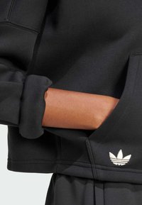 Sudadera negra con puño acanalado, con un logo blanco bordado en el bolsillo. Tela suave y diseño moderno que enfatiza la comodidad.