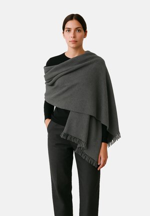 Châle en laine grise avec des bords frangés, drapé sur un haut noir, offrant une texture douce. Porté avec un pantalon noir ajusté.