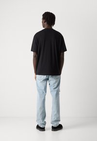 Svart bomull T-skjorte med korte ermer, sammen med lyseblå denim jeans og svarte sko, sett fra baksiden mot en ensfarget bakgrunn.