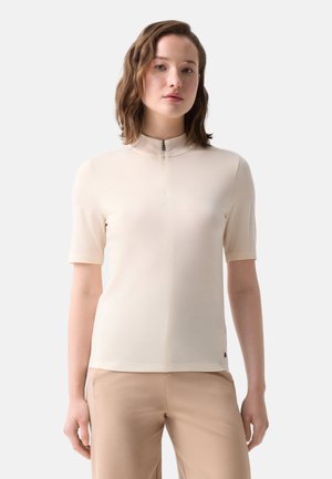 Vrouw met schouderlang haar, die een beige top met korte mouwen en een klein voorritsje draagt, en lichtbruine broek, staand tegen een effen achtergrond.