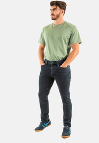 Maglietta in cotone verde a maniche corte, jeans in denim scuro, sneaker blu e grigie con dettagli blu, postura eretta, mani in tasca.