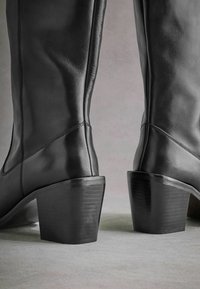 Černé kožené vysoké boty po kolena se čtveratou patou, hladkým povrchem, prošíváním a elegantním designem.