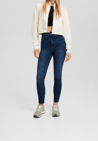 Chaqueta blanca corta con cremallera, top negro ajustado, jeans skinnies oscuros de tiro alto y zapatillas grises con acentos en blanco y negro.