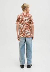 Camisa de manga corta, marrón con estampado floral y patrones blancos, combinada con jeans de color azul claro y zapatos negros. Tejido texturizado, ajuste relajado.