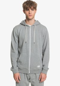 Sudadera gris con cremallera, fabricada en un material suave, que cuenta con un bolsillo tipo canguro, cordones blancos y puños acanalados. Diseño simple y casual.