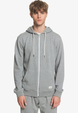 Hoodie zippé gris en tissu doux, avec une poche kangourou, des cordons blancs et des poignets côtelés. Design simple et décontracté.