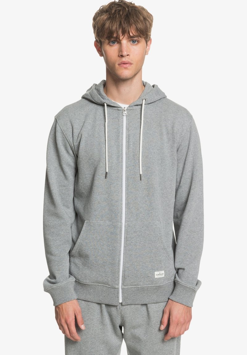 Sudadera gris con cremallera, fabricada en un material suave, que cuenta con un bolsillo tipo canguro, cordones blancos y puños acanalados. Diseño simple y casual.