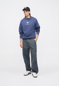 Sweatshirt azul com estampa de urso, calças cinzentas e ténis brancos. Boné às riscas a preto e branco. Tecido liso, corte relaxado, detalhes mínimos.