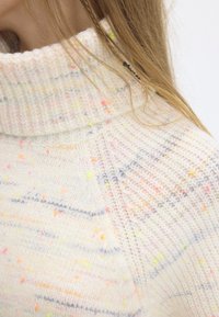 Pull couleur crème avec un col haut côtelé, parsemé de taches colorées et de fines rayures bleues, confectionné dans un matériau tricoté doux et texturé.