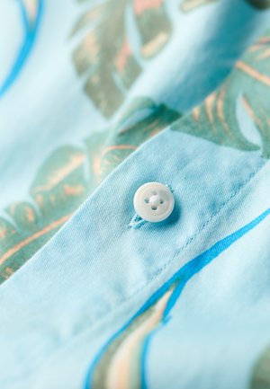 Lichtblauwe stof met een bladerenpatroon in groen en roze. Een witte knop is gestikt aan de rand, nabij de vouw van de stof.
