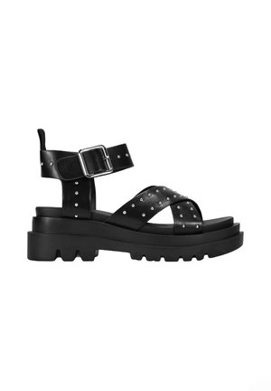 Sandal negra con detalles de remaches, correas anchas y cierre de hebilla. Suela gruesa y resistente que ofrece tracción y estabilidad.