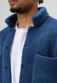 Blauwe fleecejas met een gestructureerd patroon, voorzien van een kraag, knoopsluitingen en een borstzak. Draagt over een wit t-shirt.