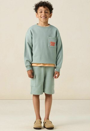 Sweatshirt vert menthe avec poche et texte, associé à un short assorti avec poches latérales, porté avec des mocassins beige.