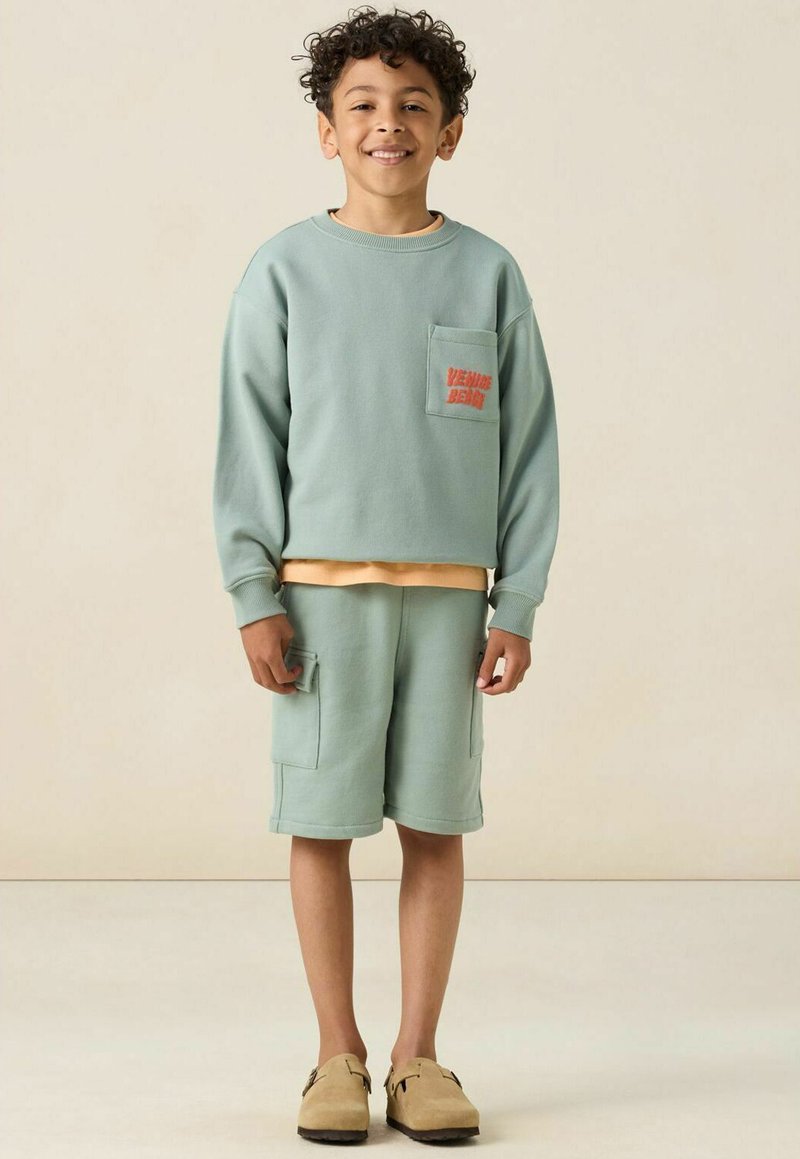 Sweatshirt vert menthe avec poche et texte, associé à un short assorti avec poches latérales, porté avec des mocassins beige.