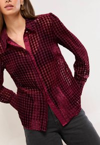 Burgundijas pogu krekls ar hounds-tooth rakstu, caurspīdīgu tekstūru un garām piedurknēm. Iespējojusies klasiskā apkakle un priekšējā kabatiņa.