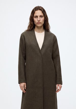 Kvinde med langt brunt hår iført en lang, mørk olivengrøn frakke over en lys beige top, stående mod en ensfarvet lys baggrund.