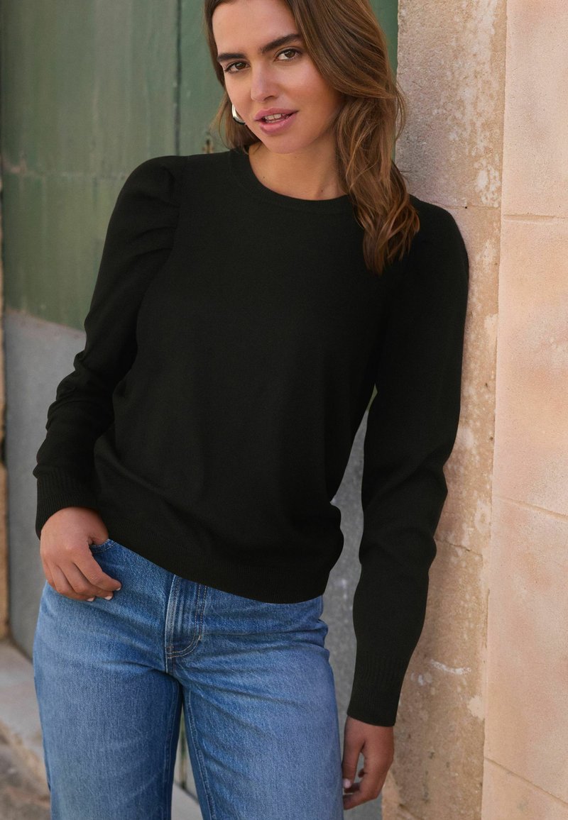 Schwarzer gestrickter Pullover mit Rundhalsausschnitt, Puffärmeln und gerippten Bündchen, kombiniert mit blauen Jeans. Strukturiertem Steinhintergrund.