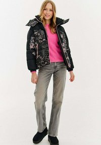 Czarna kurtka puchowa z kwiatowymi wzorami, różowymi akcentami i odpinanym kapturem, noszona na jasnoróżowym swetrze i szarych dżinsach. Czarne botki za kostkę.