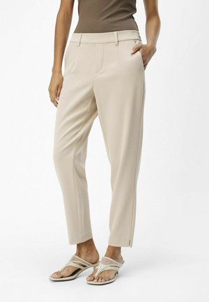 Vrouw draagt beige op maat gemaakte enkellange broek met zijzakken, gecombineerd met witte sandalen met hak en een mouwloze bruine top.