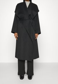 Langer schwarzer Wickelmantel aus strukturiertem Stoff mit weiten Ärmeln, einem Bindegürtel und einer fließenden Silhouette. Über einer schwarzen Hose getragen.